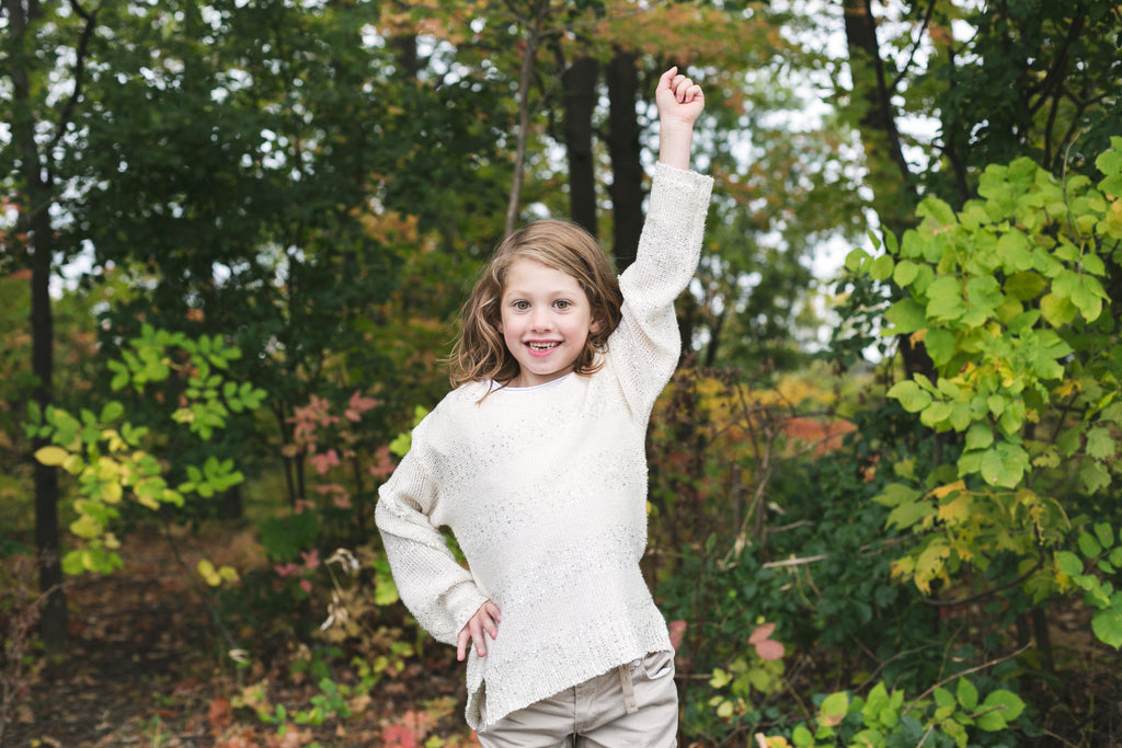 Elementary school aged girl standing in the woods wearing a white sweater. The colours of the leaves on the trees are changing for fall.