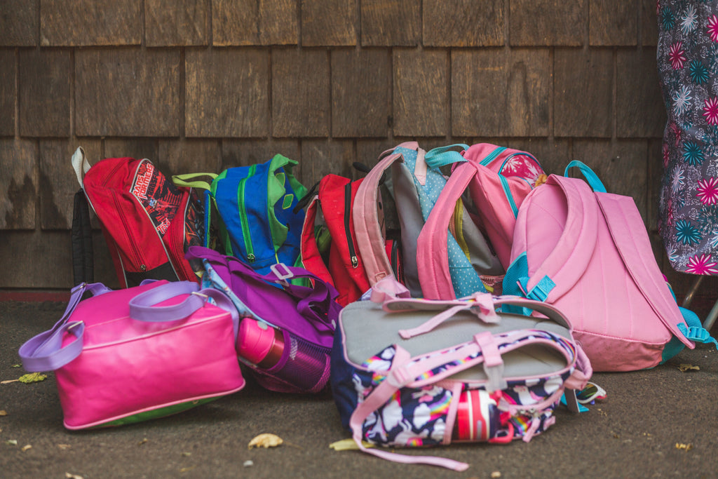 A pile of colourful childrens' backpacks sitting on the ground outside of school.
