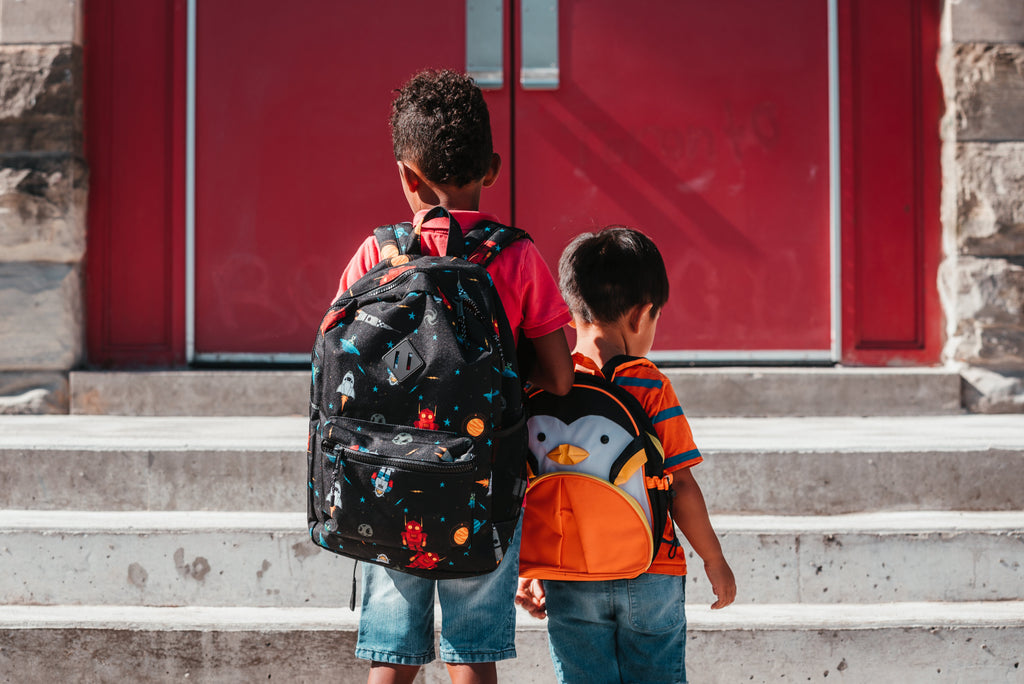 Two elementary school aged children wearing backpacks and walking toward red school doors.